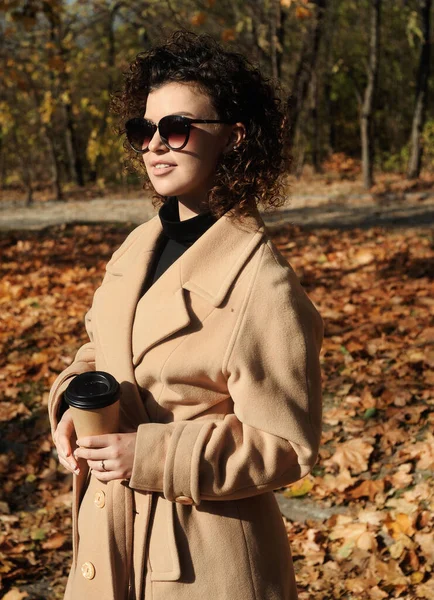 穿着时髦米黄色外套 在户外喝杯咖啡的年轻卷发女人 美丽的年轻女子在秋天的公园里喝咖啡 绿叶飘落 — 图库照片