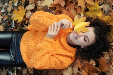 Sonbahar yaprağıyla gülümseyen genç kadın sonbahar parkında eğleniyor. Dışarıdaki yeşillikte kıvırcık saçlı mutlu bir kadının portresi..