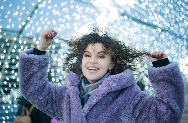 流行の色の紫色のコートで笑顔巻き毛の女性の肖像画は屋外でクリスマスライトでお祝いの背景にポーズ — ストック写真