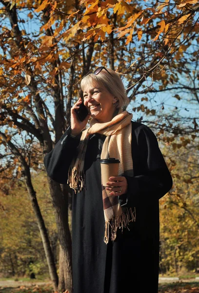  Orta yaşlı güzel bir kadın dışarıda kahve içiyor. Elinde bir fincan kahveyle sonbahar parkında yürüyen olgun bir kadın..