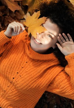 Sonbahar yaprağıyla gülümseyen genç kadın sonbahar parkında eğleniyor. Dışarıdaki yeşillikte kıvırcık saçlı mutlu bir kadının portresi..