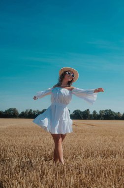 portrait of young curly haired woman in white dress with a white sunglasses posing in the wheat field