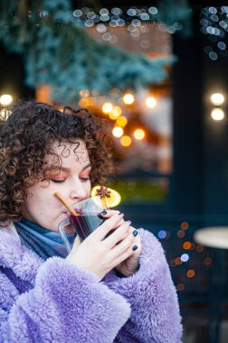 Gülümseyen kıvırcık saçlı, modaya uygun menekşe ceketli kadın açık havada sıcak şarap içiyor. 