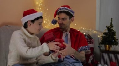 Noel Tatili 'nde iki adam ellerinde hediyelerle kanepede oturuyor. Noel Baba şapkasıyla. Noel zamanı. Mutluluk