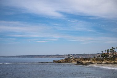 La Jolla, California, ABD - 3 Ekim 2021: Mavi bulutların altında, mavi okyanusun üzerindeki kahverengi kayalıklarda çocuk havuzu hamamı. Palmiyeli kayalar ve plajlar.