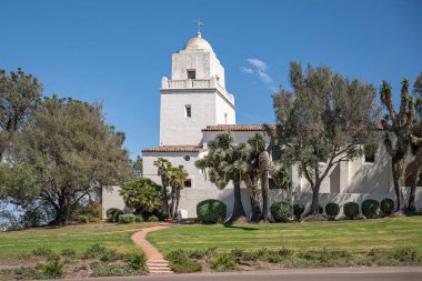 San Diego, California, ABD - 3 Ekim 2021: Junipero Serra Müzesi kulesi tepedeki mavi gökyüzüne karşı beyaz bina ve önde yeşil yapraklar