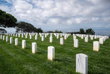San Diego, California, USA - October 5, 2021: Fort Rosecrans National Cemetery. Rows of white tombstones on green grass in front of Pacific Ocean under heavy blue cloudscape. Green foliage. clipart
