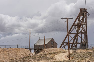 Tonopah, Nevada, ABD - 18 Mayıs 2011: Tarihi Madencilik Parkı. Kuru bej çöl zemininde daha yüksek binalarla ahşap maden kuyusu kulesi ve yukarıdaki ağır gri bulutların karışımı..