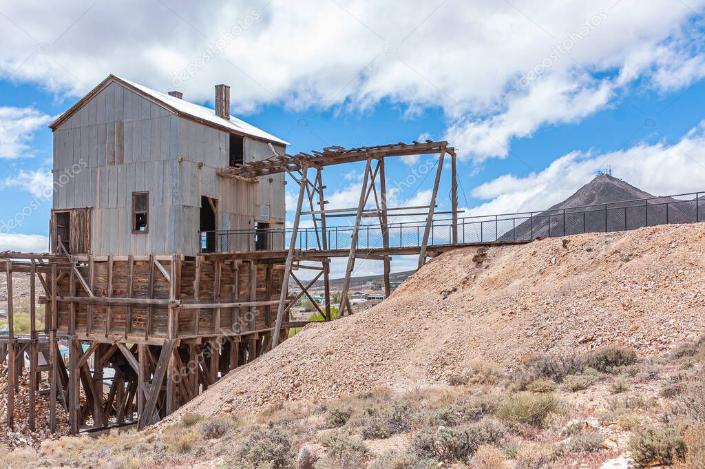 Tonopah, Nevada, US - May 18, 2011: Historic Mining Park. Gray corrugated metal sheet building above pit and beige-brown heap of stripping or waste rock under blue cloudscape.