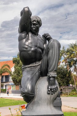 San Diego, California, USA - October 4, 2021: Closeup on black stone Morning statue by Donald Hord on pedestal in front of Wild blue-gray cloudscape,. Buildings of boardwalk and green foliage.