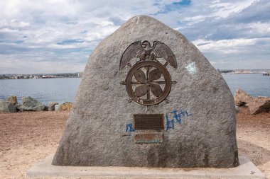 San Diego, California, USA - October 4, 2021: Merchant Seamen Memorial Statue at Embarcadero Marina Park is gray boulder with metal edge of eagle and boat steering wheel. Blue cloudscape.