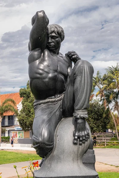 San Diego, California, USA - October 4, 2021: Closeup on black stone Morning statue by Donald Hord on pedestal in front of Wild blue-gray cloudscape,. Buildings of boardwalk and green foliage.