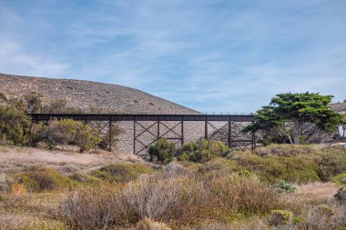Lompoc, California, USA - November 19, 3021: Jalama Beach. Dry green-yellow landscape along creek under brown metal-wooden bridge of the Pacific Railroad under blue cloudscape.