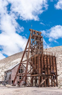 Tonopah, Nevada, ABD - 19 Mayıs 2011: Koyu kahverengi ahşap kulenin kapatılması ve terk edilmiş madenlerin bej çöl zemininde mavi bulutların altında dökülmesi.