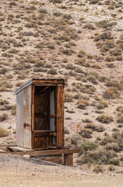 Tonopah, Nevada, ABD - 19 Mayıs 2011: Terk edilmiş madendeki kuru bej çöl zeminine kurulmuş açık kahverengi ahşap tuvaletin kapatılması.