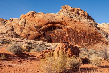 Overton, Nevada, ABD - 11 Aralık 2010: Ateş Vadisi. Mavi gökyüzünün altındaki dağ sırasının kızıl kayalıklarındaki doğal kemer manzarası. Önünde kuru çalılar olan çöl tabanı..