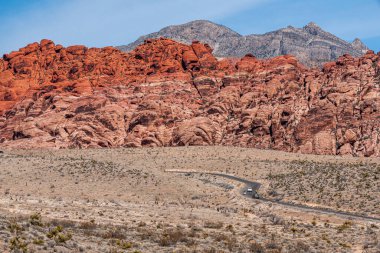 Las Vegas, Nevada, USA - February 23, 2010: Red Rock Canyon Conservation Area. Wide landscape shows road with cars meandering in dry desert valley. Red rock wall under blue sky. clipart