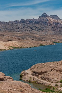 Nelson, Nevada, ABD - 21 Mayıs 2011: Mavi Colorado Nehri üzerindeki küçük tekne, dağlarla, tepelerle ve mavi bulutların altındaki bazı yeşil bitkilerle birlikte kuru çöl arazisini kesiyor..
