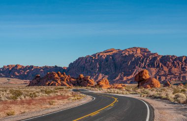 Overton, Nevada, ABD - 24 Şubat 2010: Ateş Vadisi. Mavi gökyüzünün altında gün batımı manzarası. Siyah asfalt yolu. Kırmızı renkli dağlar ve kayalar ile kuru çölde geziniyor..