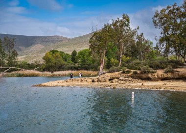 Winchester, CA, USA - 11 Nisan 2022: Skinner Gölü. Kumlu sahil şeridi ve mavi suların arkasındaki ağaçlar. Aile yolu arkada. Ufuktaki tepelerde yeşil yapraklar, hepsi mavi bulutların altında.