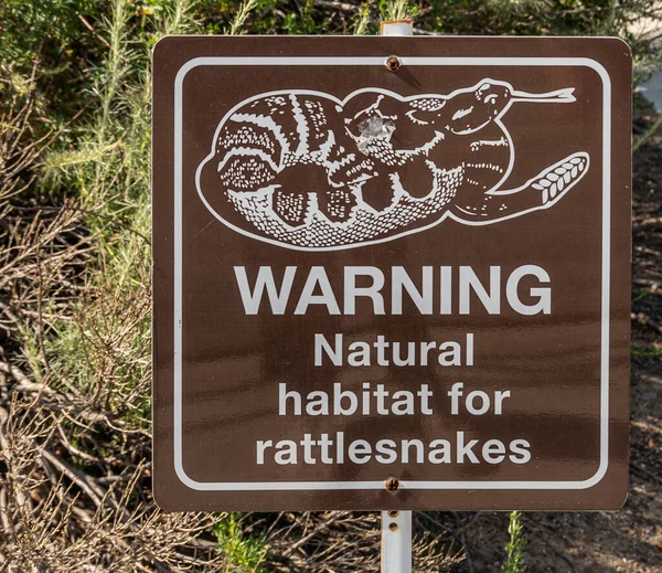 Simi Valley, California, USA - April 27, 2022: Closeup of warning sign about presence of rattlesnakes in their natural habitat. White image and letters on dark brown square plate. Green foliage in back.