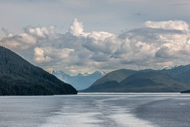 Pasifik Sahil Hattı, Alaska, ABD - 16 Temmuz 2011: Açık mavi gökyüzünde ağır bulutların altında Karlı Kanada Rocky Dağları. Mavi kıyı sularında yeşil ormanlık tepelerle çevrili gemi izleri.. 