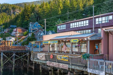 Ketchikan, Alaska, USA - July 17, 2011: Retail businesses built on wharf along Water Street featuring coffee shop, fishing charters, snacks, laundry services, and more. Green forest in back. clipart
