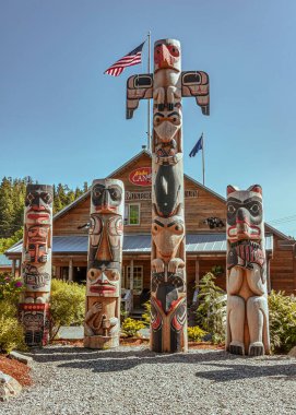 Ketchikan, Alaska, ABD - 17 Temmuz 2011: Yağmur Ormanları Sığınağı. Kahverengi binanın önündeki 4 renkli totem direğinin portresi. Alaska Yağmur Ormanları Sığınağı. Mavi gökyüzünün altında. Çevrede yeşil yapraklar.