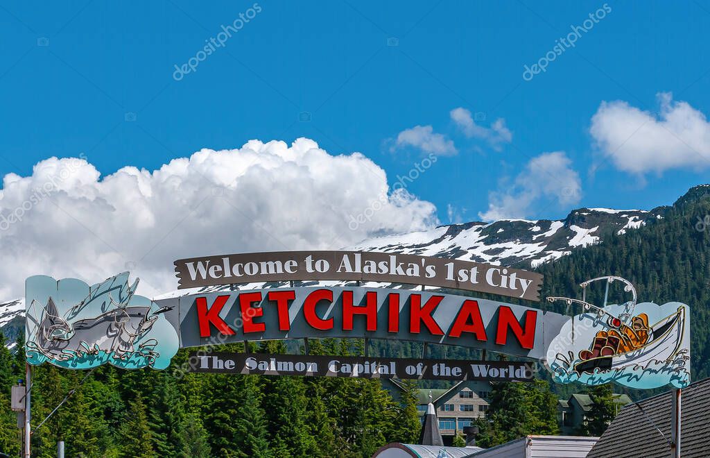 Ketchikan, Alaska, USA - July 17, 2011: Large free standing colorful Welcome sign to the Salmon Capital of the World under blue cloudscape and snow covered mountain on horizon. Green foliage.