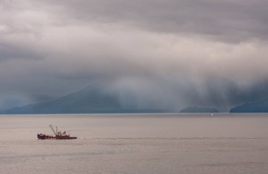 Hoonah, Alaska, ABD - 18 Temmuz 2011: Sisli bulutlar ufuk üzerinde kısmen görünür orman dağları üzerinde yağmura dönüşürken derin marroon okyanus kızı balıkçı teknesi kahverengi suda hareket ediyor.