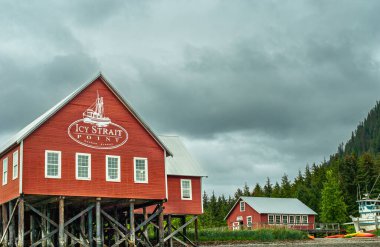 Hoonah, Alaska, USA - July 18, 2011: Red Ice Strait Point buildings with emblem at deep water harbor under gray cloudscape. Green forest in back and up hill. clipart