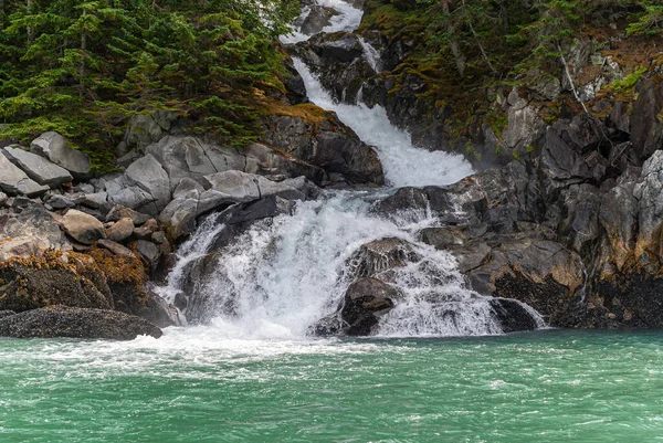 Skagway, Alaska, USA - July 20, 2011: Taiya Inlet above Chilkoot Inlet. White surf of waterfall reaches green ocean water over brown-gray rocky shoreline with green foliage higher up.
