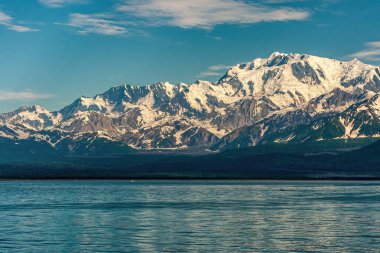 Hüsran Körfezi, Alaska, ABD - 21 Temmuz 2011: Yakuta Körfezi, güneş, mavi bulutların altında okyanus kıyısındaki dağlara kar yağdırıyor. Önde mavi okyanus suyu var. Ufuktaki ormanlık kıyı şeridi
