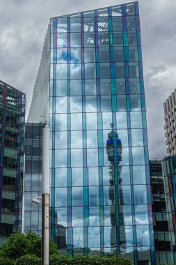 London, Great Britain - July 3, 2022: Tall slender BT tower reflected in blueish glass facade of Regent's Place office building. Green foliage at bottom and gray cloudscape.