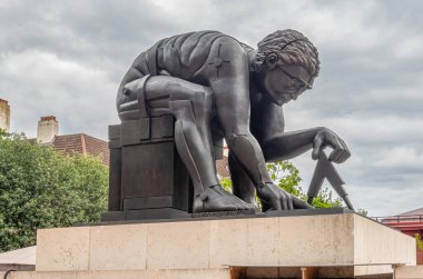 London, Great Britain - July 3, 2022: Closeup of gray Newton Statue on pedestal in front yard of British Library and Knowledge Center under gray cloudscape. Some green foliage
