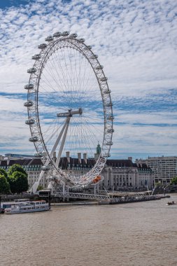 London, UK - July 4, 2022: White London Eye against blue cloudscape. Brown water Thames River in front. Cityscape of multi-story buildings behind.