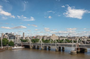 London, UK - July 4, 2022: Golden Jubilee bridges on both sides of Hungerford Bridge spanning brown water Thame river under blue cloudscape. Charing Cross railway station and cityscape.
