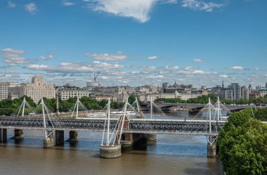 London, UK - July 4, 2022: Wide cityscape with Hungerford and Waterloo bridges over brown water Thames River under blue cloudscape. Green foliage adds color.