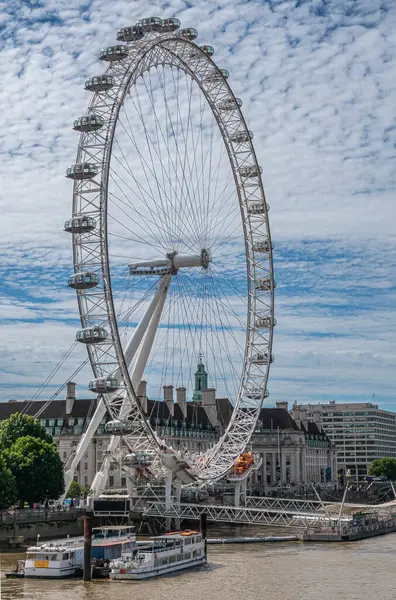 London, UK - July 4, 2022: Closeup of White London Eye against blue cloudscape. Brown water Thames River in front. Cityscape of multi-story buildings behind.