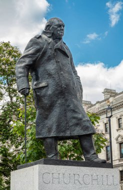 London, UK - July 4, 2022: Parliament Square Gardens. Closeup of Bronze statue of Winston Spencer Churchill on gray pedestal under blue cloudscape. Green foliage in back.