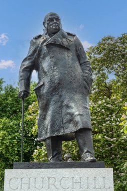 London, UK - July 4, 2022: Parliament Square Gardens. Frontal Closeup of Bronze statue of Winston Spencer Churchill on gray pedestal under blue cloudscape. Green foliage in back.