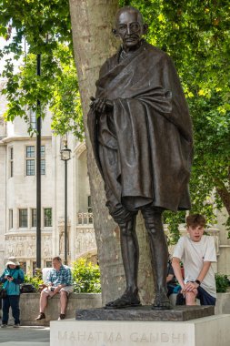 London, UK - July 4, 2022: Parliament Square Gardens. Bronze statue of Mahatma Gandhi, Campaigner for Indian Independence, backed by green foliage. People around.