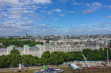 London, UK - July 4, 2022: Seen from London Eye. New and old Whitehall buildings under blue cloudscape with construction work and boats on brown water Thames river. Green foliage belt