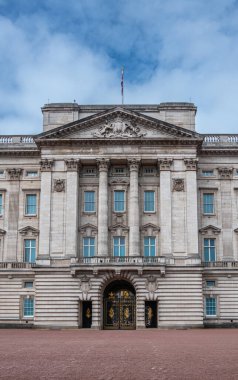 London, England, UK - July 6, 2022: Central columned part of beige-gray stone of Buckingham Palace facade under blue cloudscape. Pediment above under British flag. Black-Gold entrance doors