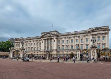 London, England, UK - July 6, 2022: Buckingham Palace. Lots of pedestrians in front of beige stone building and black fence with golden details under blueish cloudscape.