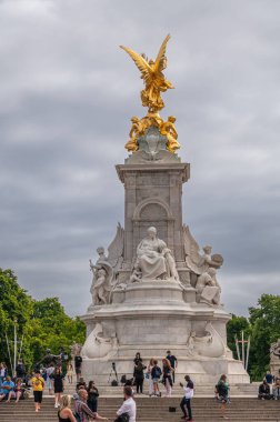 London, England, UK - July 6, 2022: Victoria Memorial with golden Victory statue. People on steps below motherhood statue under gray cloudscape. Green foliage and clothing add color