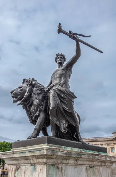 London, England, UK - July 6, 2022: Victoria Memorial. Closeup of Black bronze Progress statue of lion, youth with flaming torch under blue cloudscape. Some green foliage