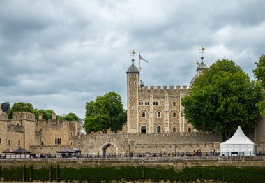 London, England, UK - July 6, 2022: Tower of London. White tower palace building seen over ramparts from Thames River under gray cloudscape. People on quay.