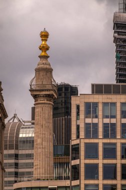 London, England, UK - July 6, 2022: From Thames River. Monument to the Great Fire of London features golden flame symbol on top of thick tall beige stone column against gray sky. Office buildings around