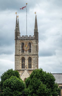 London, England, UK - July 6, 2022: From Thames River. Gray stone tower of Southwark Cathedral with 4 spires, 1 tall flag on top against gray cloudscape. Green foliage up front.